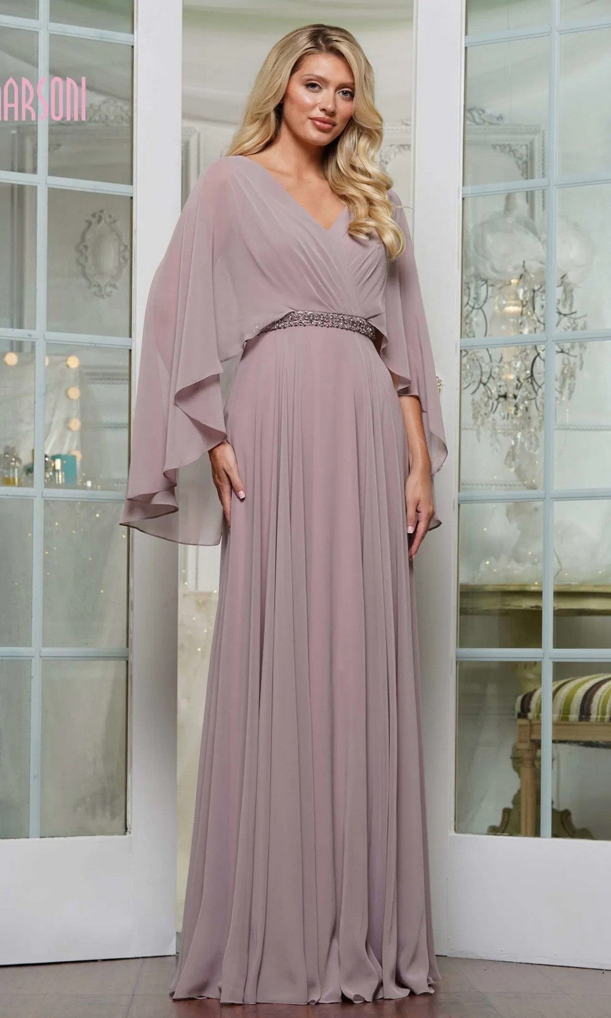 A woman poses indoors in front of glass double doors, wearing the Marsoni MV1350 Long Formal Dress in dusty rose chiffon, featuring flowing sleeves and an embellished waistbelt.